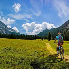 Dvoje biciklista voze planinskim putem kroz prirodu i zelene livade tokom sunčanog dana u okviru outdoor aktivnosti i rekreacije na otvorenom.