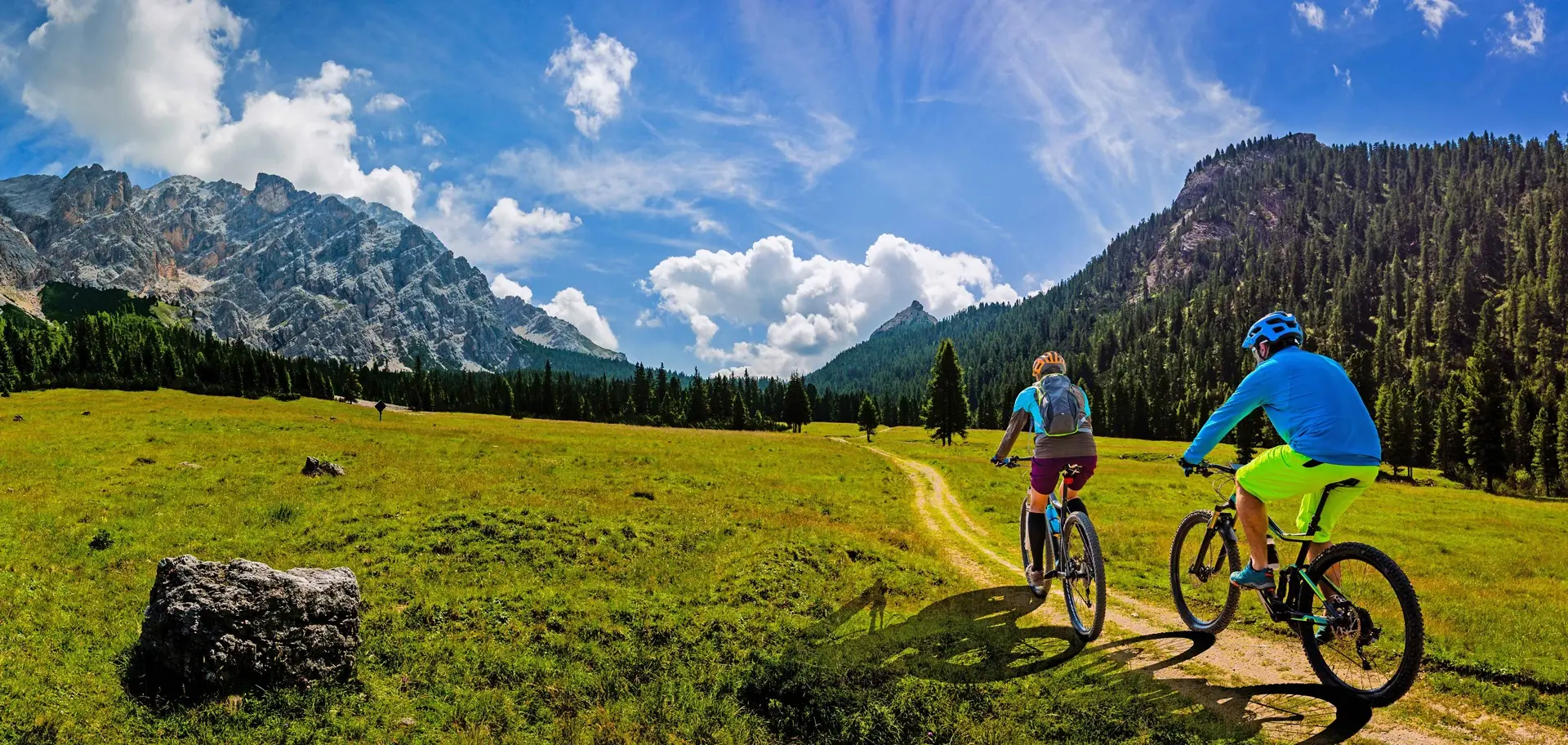 Dvoje biciklista voze planinskim putem kroz prirodu i zelene livade tokom sunčanog dana u okviru outdoor aktivnosti i rekreacije na otvorenom.